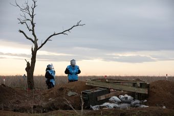 Bildet viser et åpent landskap med en vidstrakt, brunaktig eng under en grå og litt dyster himmel. Til venstre står et ensomt, bladløst tre. Midt i bildet står to personer iført blå vernedrakter med beskyttelsesutstyr på hodet. 
