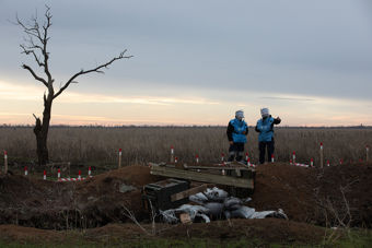 Fotografi av et landskap med en enslig, bladløs tre til venstre og to personer i blå beskyttelsesvester til høyre. De står ved et område markert med rødt og hvitt, sannsynligvis et mineryddingsområde. Bakgrunnen viser en vidstrakt eng under en overskyet himmel.