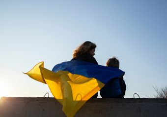 To personer sitter side om side på en murkant, sett bakfra mot en klar, blå himmel. Den ene personen har et stort ukrainsk flagg, blått og gult, drapert rundt skuldrene. Flagget vaier i vinden og dekker begge personene, noe som skaper et inntrykk av samhold og beskyttelse. Solen er lav på himmelen, og lyset gir flagget et varmt, gyllent skjær. 