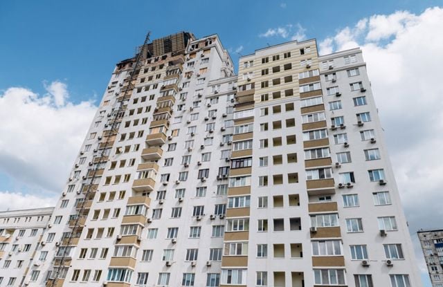The image shows a tall residential apartment building, likely more than fifteen stories high. The building is painted mostly white, with some beige and light brown accents around the windows and balconies. There are numerous windows and small balconies on each floor, and many of the windows have air conditioning units installed beneath them.