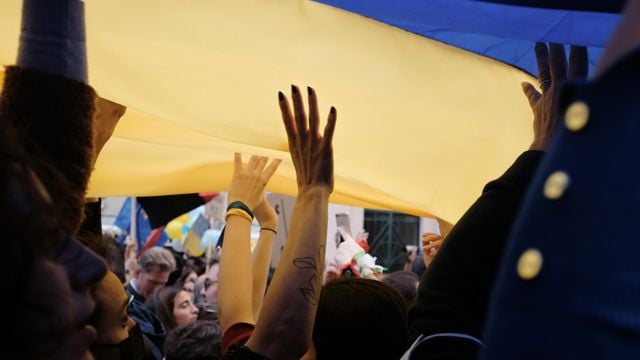 og denne Bildet viser mennesker som deltar i en folkemengde, med armene løftet mot et stort blått og gult flagg, som ligner Ukrainas flagg. Stemningen er energisk og engasjert, med mange personer samlet i en tett gruppe.
