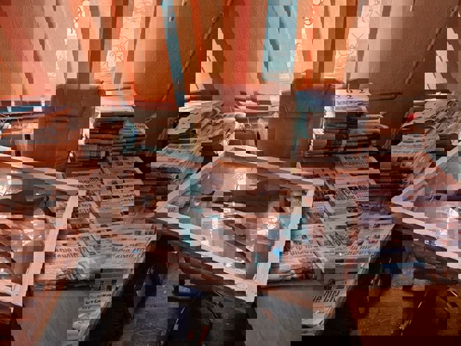 Bildet viser et kontor med et skrivebord og en kontorstol. På bordet og rundt stolen ligger store bunker med aviser, noen tydelig merket med årstallene 2022 og 2023. Midt på stolen og bordet ligger en stor, hvit vindusramme med knust glass. Bakgrunnen har vinduer med oransje gardiner som slipper inn lys, og det er en generell følelse av rot og uorden i rommet.