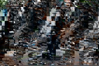 A woman in dark clothing stands among debris and damaged buildings, taking a photo with her phone.