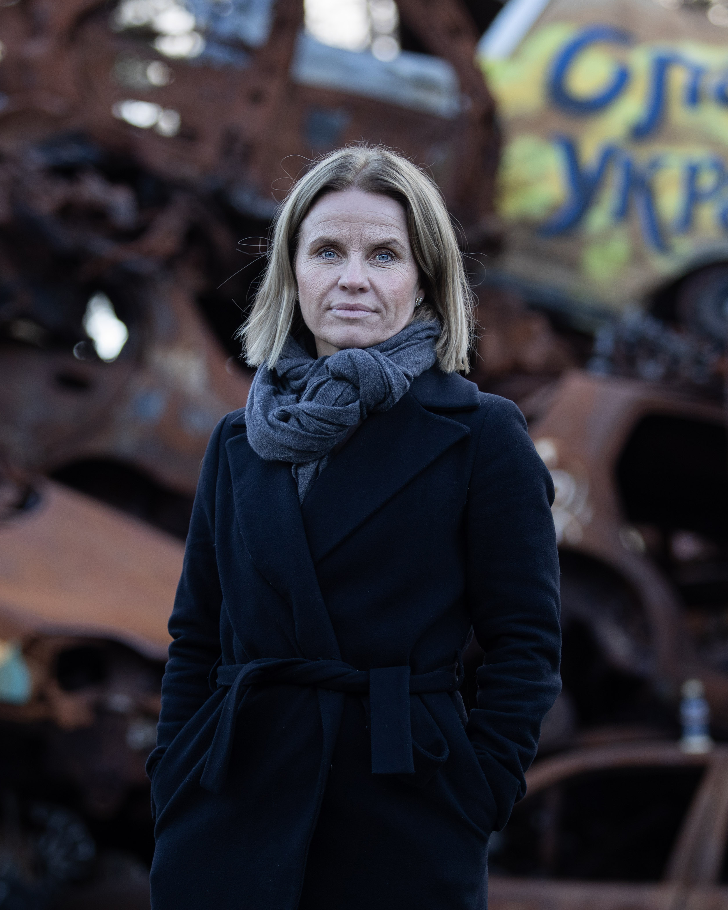 The image shows a woman with light brown hair, wearing a dark coat and gray scarf, standing in front of a pile of rusted, damaged cars. One car in the background has graffiti that reads "Ukraine.