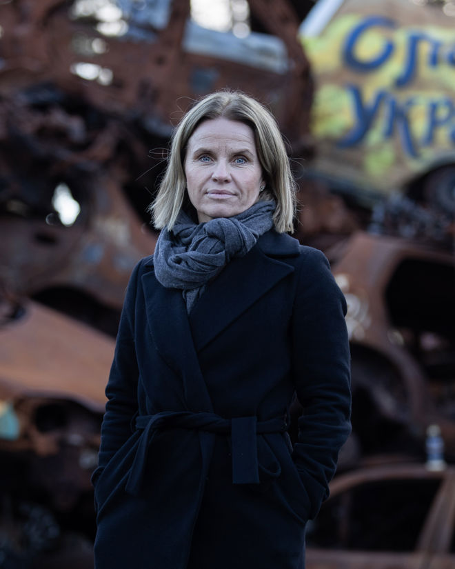 The image shows a woman with light brown hair, wearing a dark coat and gray scarf, standing in front of a pile of rusted, damaged cars. One car in the background has graffiti that reads "Ukraine.