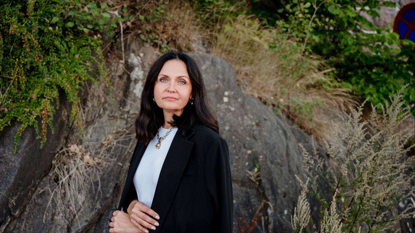A woman with shoulder-length dark hair is standing outdoors in front of a rocky wall surrounded by greenery and wild plants. She is wearing a white top, a black blazer, and a necklace. The setting feels natural and calm, with plants and rocks forming the background.