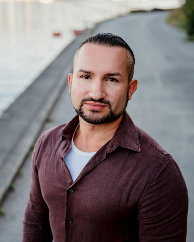 Bildet viser en mann stående utendørs på en vei ved vannkanten. Han har på seg en mørk rødbrun skjorte over en lys t-skjorte. Bakgrunnen er uskarp, men du kan se asfalt, vann og litt grønt langs kanten. Stemningen er rolig, og det ser ut til å være dagslys.