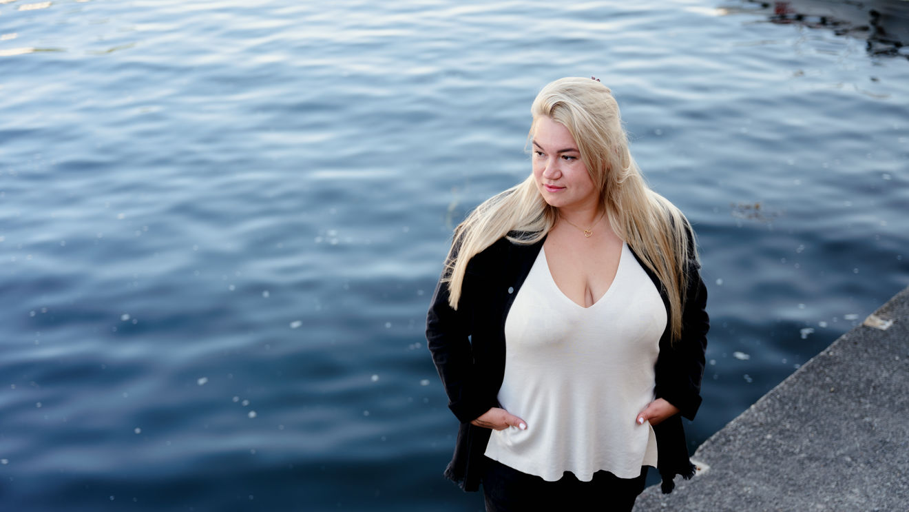 The image shows a woman with long blonde hair standing on the edge of a dock or waterfront.