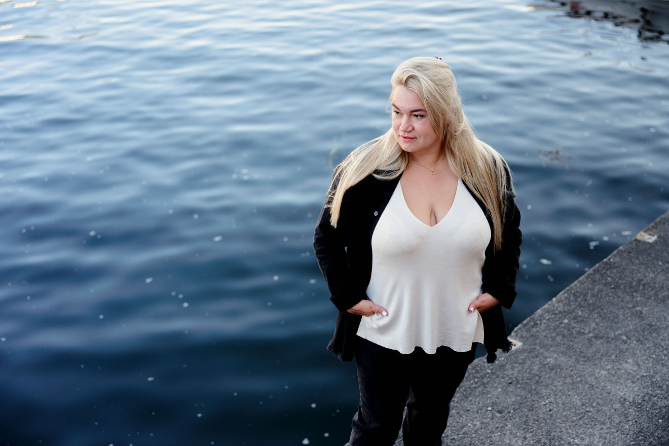 Bildet viser en kvinne med langt lyst hår som står ved kanten av en brygge eller kai, med vannet rett bak seg.