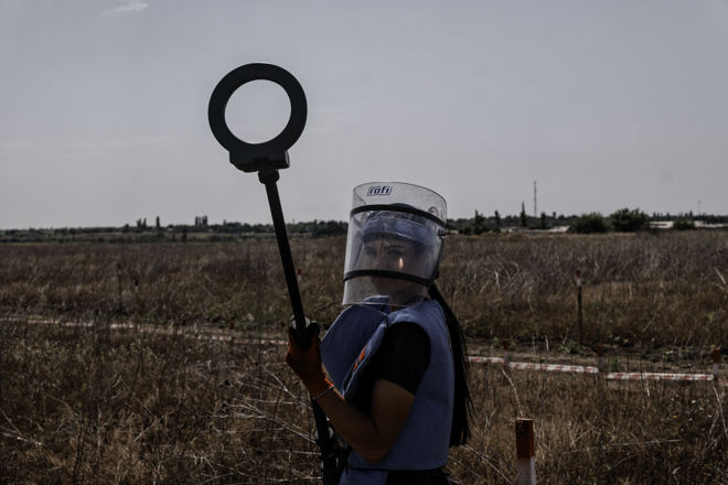 Kvinne som holder en minerydder ser i kameraet. Tomt og tørt landskap bak henne.