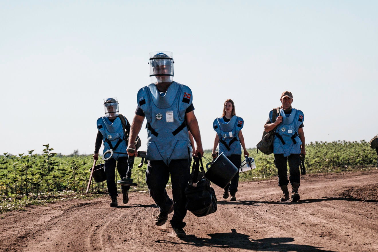 Four people in blue vests are walking on a dusty road.