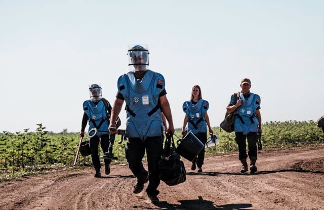 Bildet viser en kvinne i beskyttelsesutstyr, inkludert en blå vest og ansiktsskjerm, stående i et åpent landskap med tørre planter. Personen holder et verktøy som kan være en metalldetektor, noe som antyder at de utfører arbeid relatert til sikkerhet eller minerydding.
