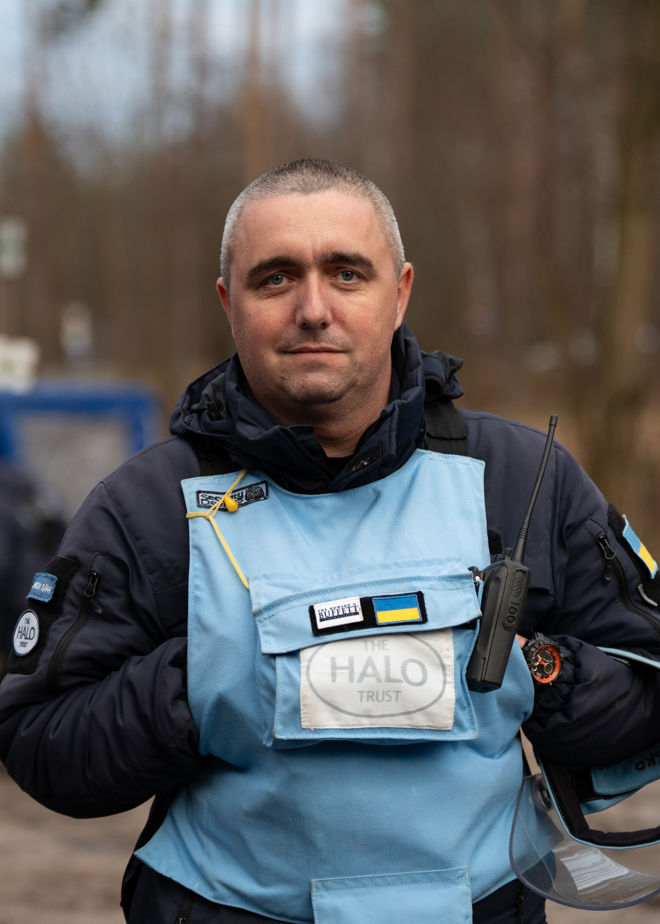 En mann iført blå beskyttelsesvest med «HALO Trust»-logo og ukrainsk flagg holder en radio og et visir, utendørs i et skogsområde.