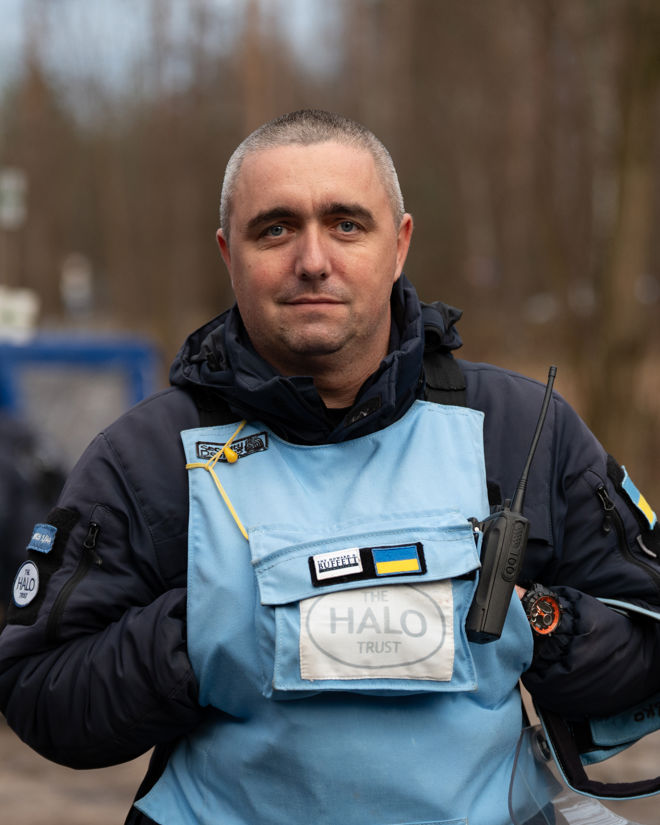 En mann iført blå beskyttelsesvest med «HALO Trust»-logo og ukrainsk flagg holder en radio og et visir, utendørs i et skogsområde.