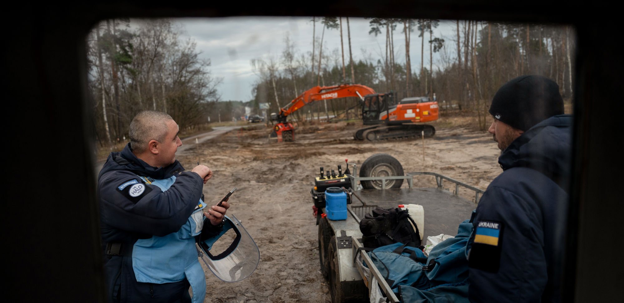 To menn i arbeidsklær står ved en tilhenger med utstyr på en gjørmete byggeplass, med en gravemaskin i bakgrunnen.
