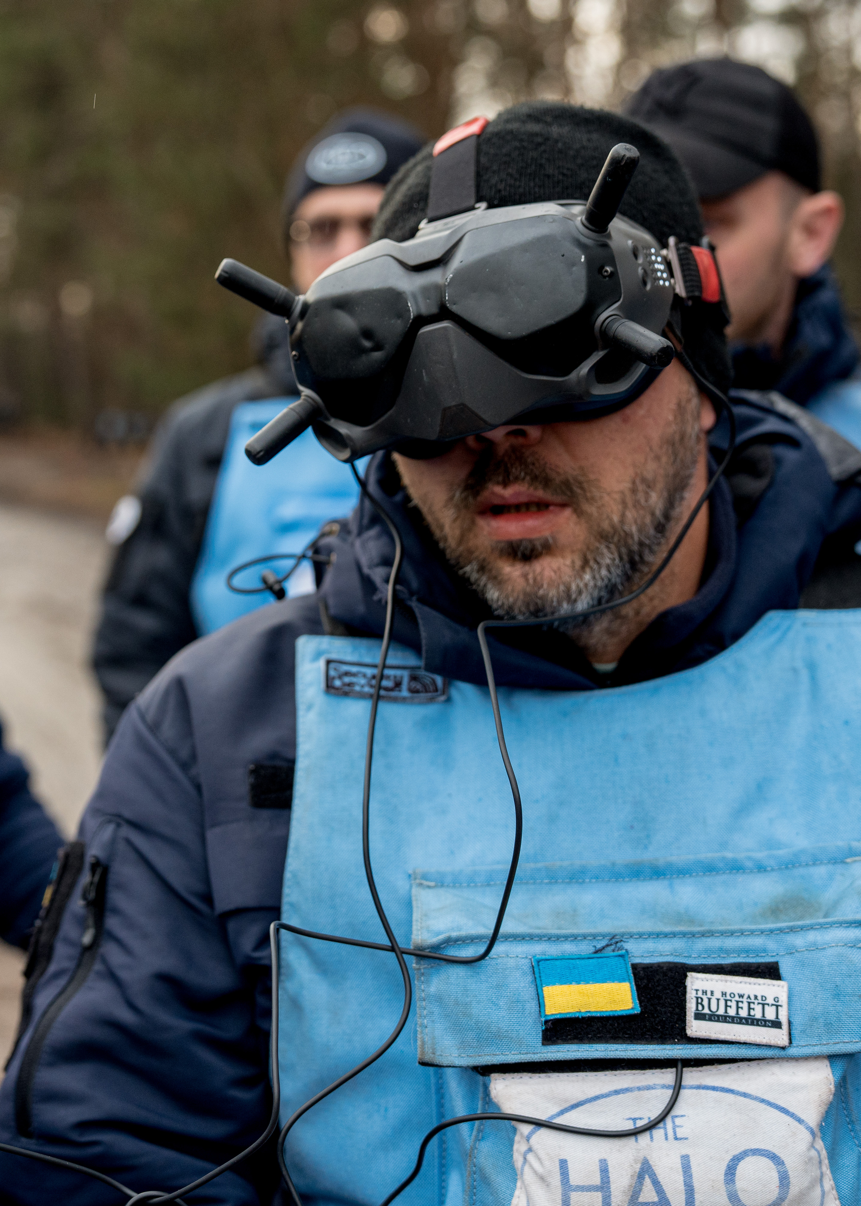 En person iført blå beskyttelsesvest og mørk lue har på seg VR-briller med ledninger, utendørs i et skogsområde.