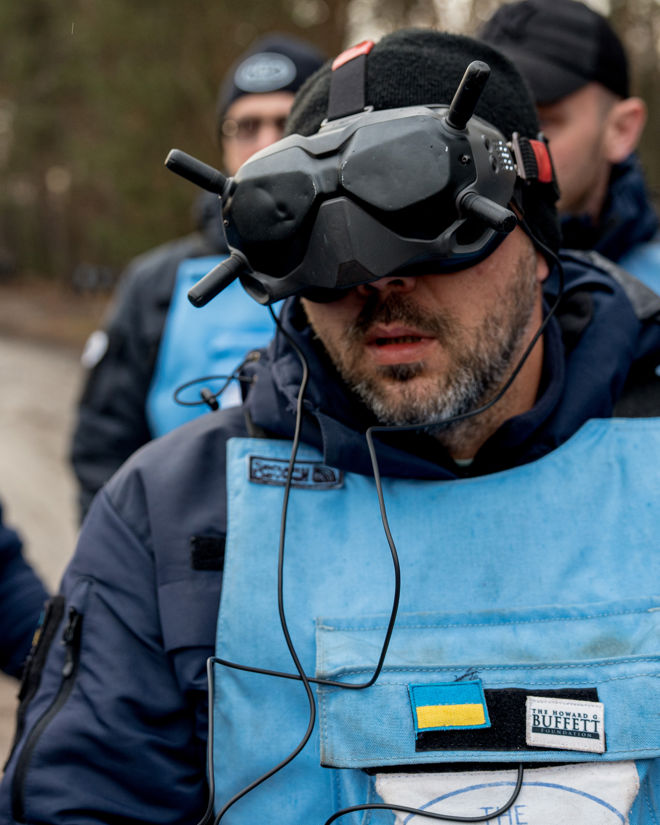 En person iført blå beskyttelsesvest og mørk lue har på seg VR-briller med ledninger, utendørs i et skogsområde.
