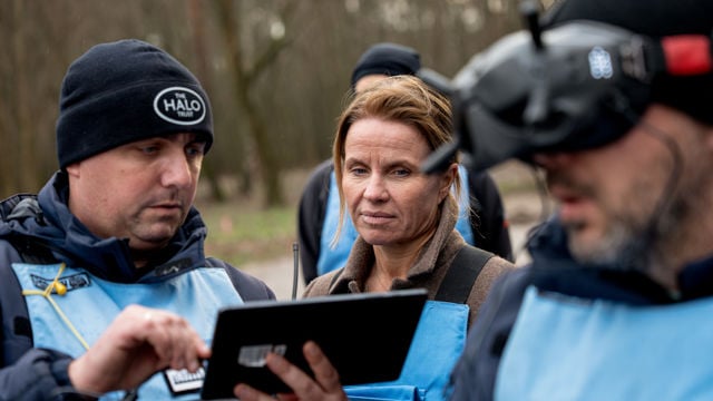Tre personer i yttertøy står tett sammen utendørs og ser på et nettbrett som en mann holder i hendene.