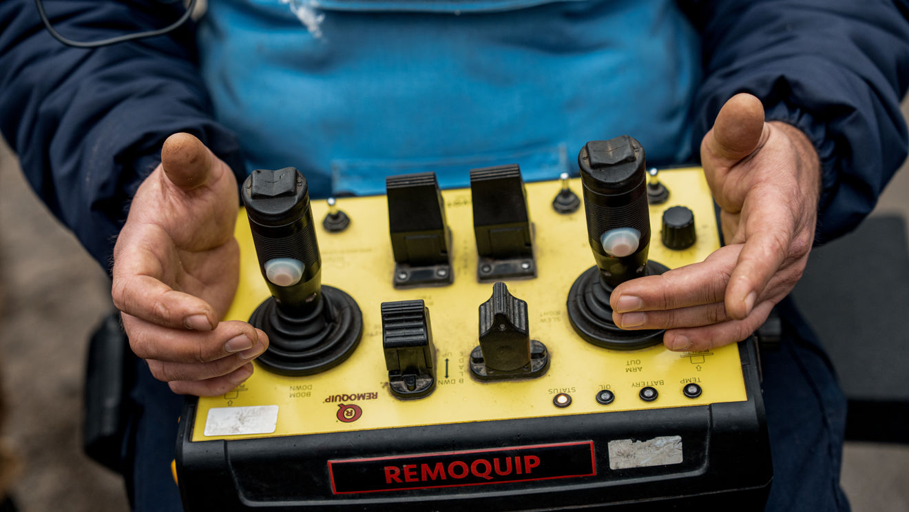 En person holder en gul fjernkontroll med to styrespaker og flere brytere, merket «Remoquip».