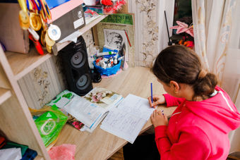 Bildet viser en ung person som sitter ved et skrivebord og gjør skolearbeid. Personen har mørkt hår satt opp i en hestehale og har på seg en rosa genser. På bordet ligger det åpne skrivebøker og papirer, og personen skriver med en blå penn. Runt skrivebordet er det mange små ting: en hylle med medaljer og bånd henger til venstre, og det står en svart høyttaler på bordet. 