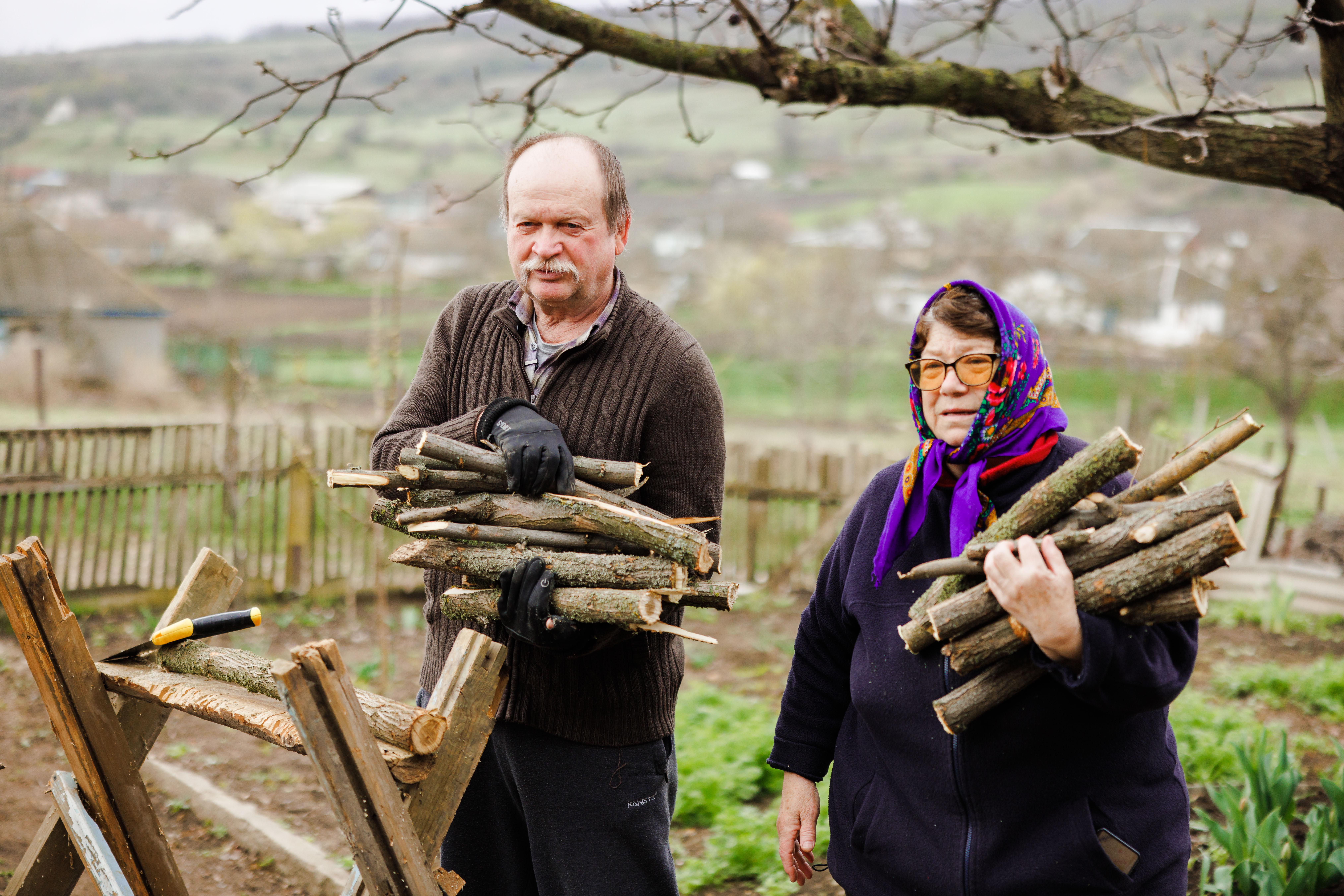 An elderly man and woman are standing outside, holding sticks for firewood.
