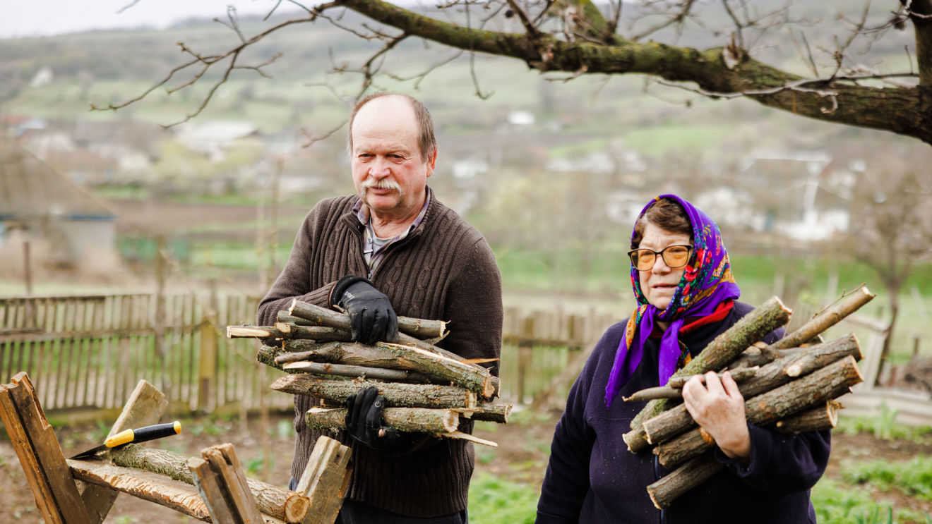 An elderly man and woman are standing outside, holding sticks for firewood.