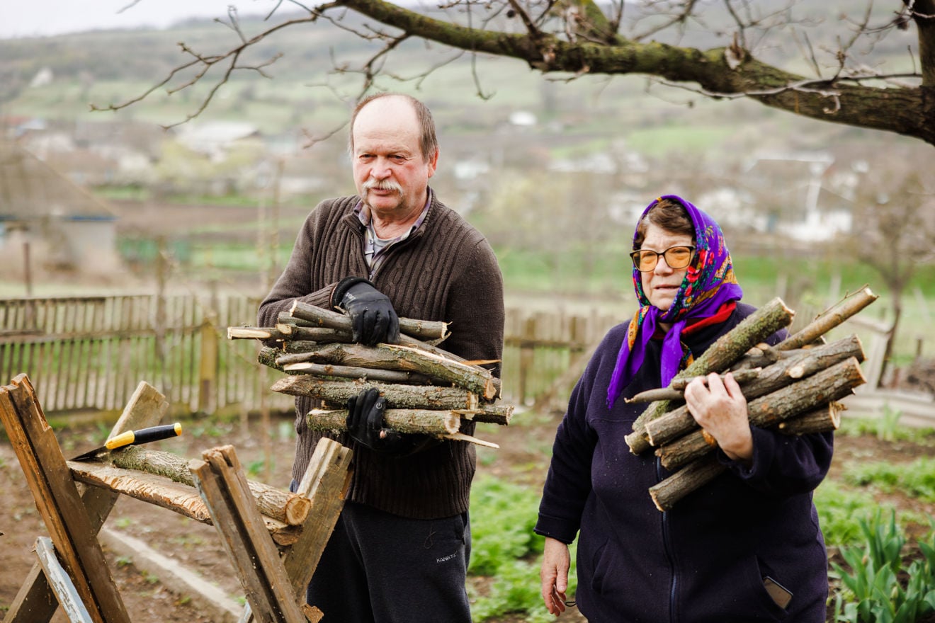 An elderly man and woman are standing outside, holding sticks for firewood.