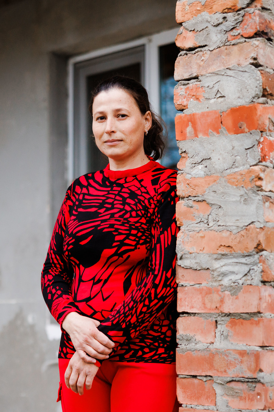 A woman in a red sweater is standing outside a house and looking at the camera.