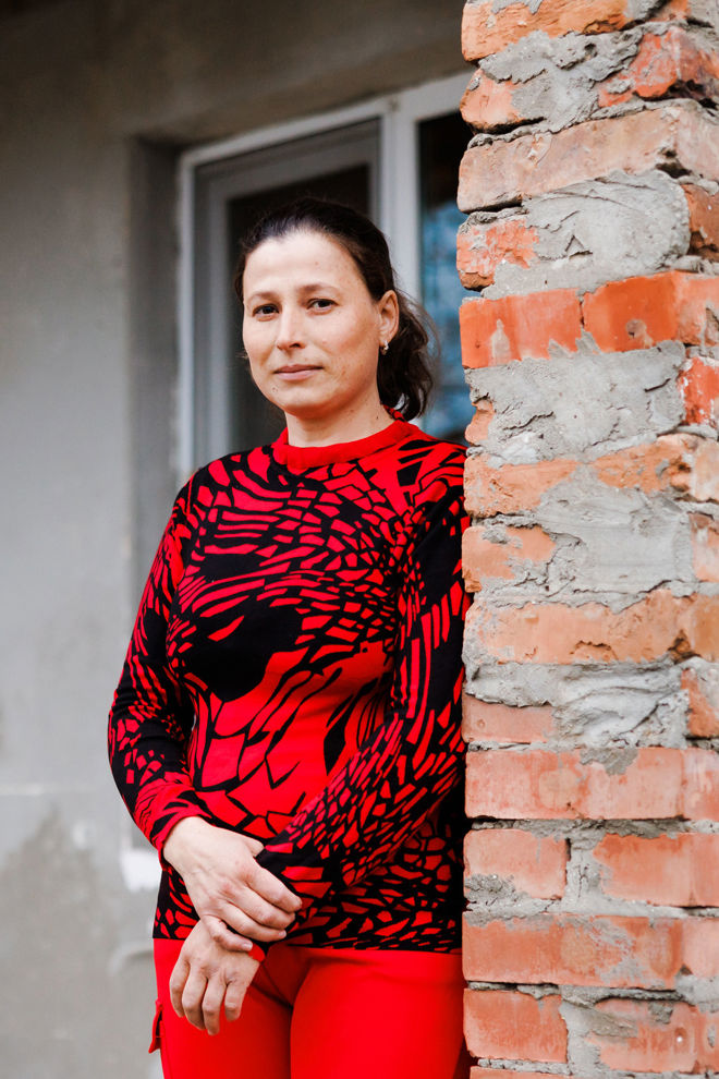 Kvinne med rød genser og mørkt hår står utenfor et hus og ser i kameraet.