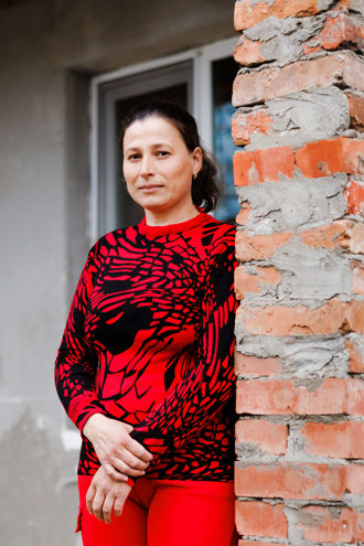 A woman in a red sweater is standing outside a house and looking at the camera.