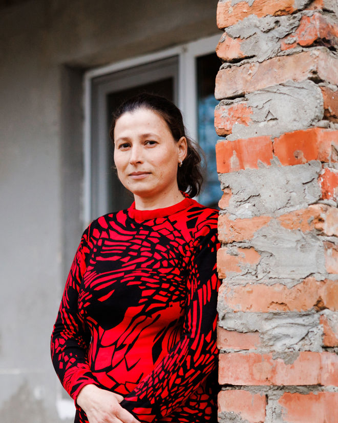 A woman in a red sweater is standing outside a house and looking at the camera.