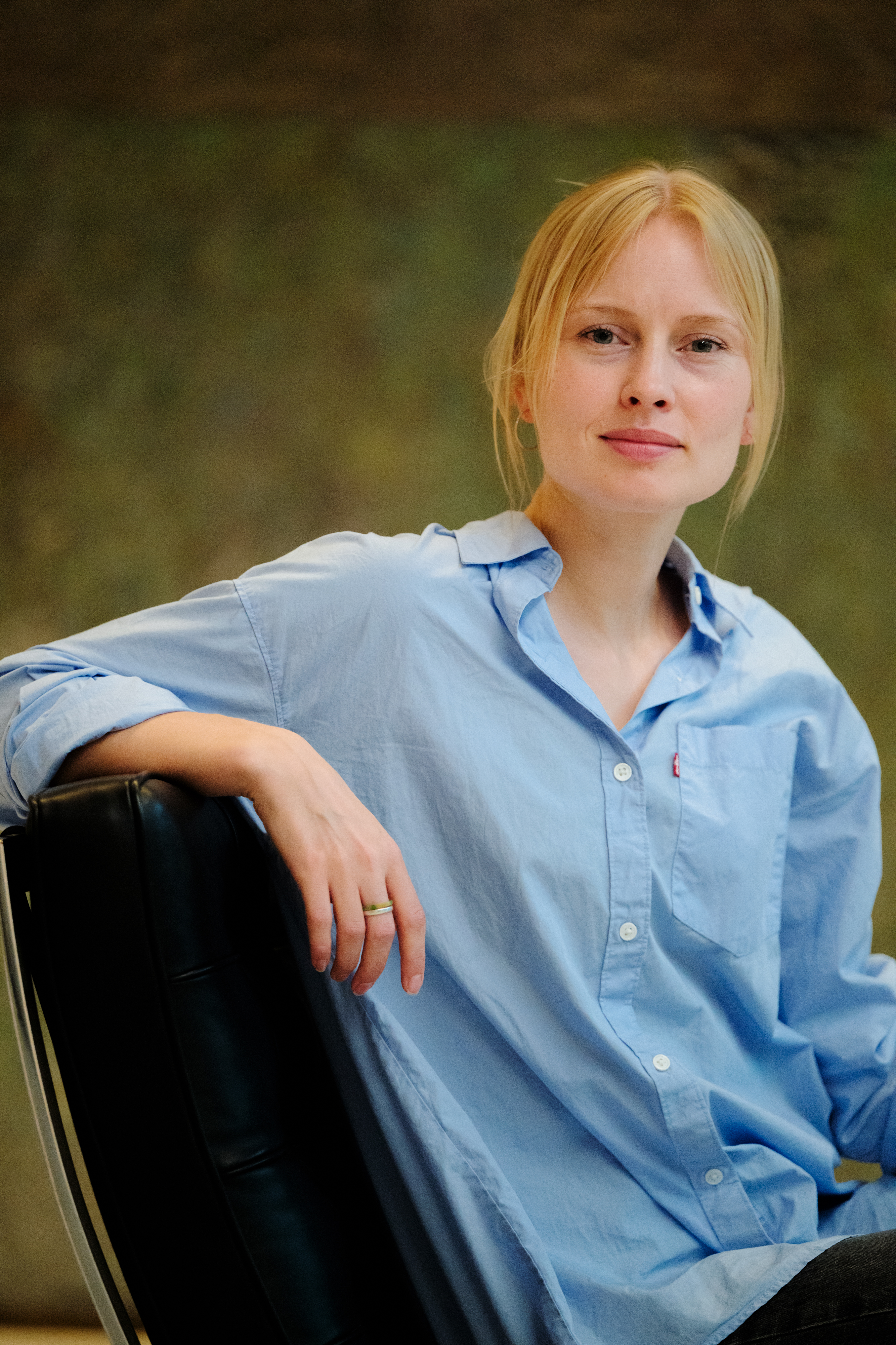 A woman is sitting in a black leather chair, wearing a light blue button-up shirt with rolled-up sleeves. The background is softly blurred in earthy tones, giving the image a calm and relaxed atmosphere. The individual has blond hair tied back and wears a simple ring on one finger. 