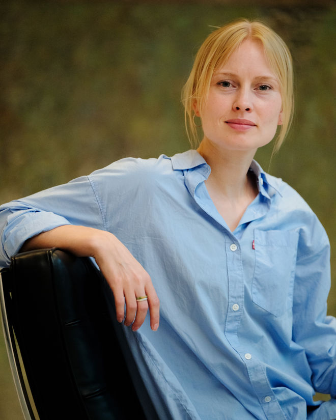 A woman is sitting in a black leather chair, wearing a light blue button-up shirt with rolled-up sleeves. The background is softly blurred in earthy tones, giving the image a calm and relaxed atmosphere. The individual has blond hair tied back and wears a simple ring on one finger.