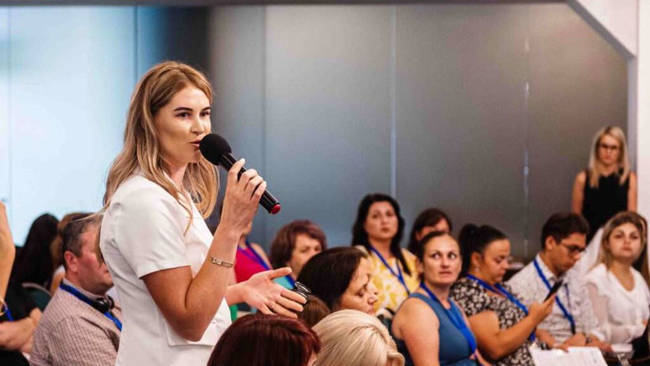 A woman in a white outfit is speaking into a microphone in front of a seated group at a conference or seminar. The audience appears attentive, with some people holding notebooks and wearing name tags. The setting is professional and focused.