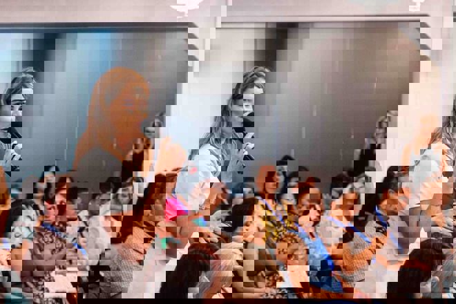 Dette bildet viser en gruppe mennesker samlet i et rom, antakelig til et møte, konferanse eller seminar. En kvinne i hvit kortermet bluse står midt i bildet og snakker i en mikrofon, mens hun vender seg mot resten av gruppen. Hun ser ut til å delta aktivt, kanskje hun stiller et spørsmål eller gir en kommentar.