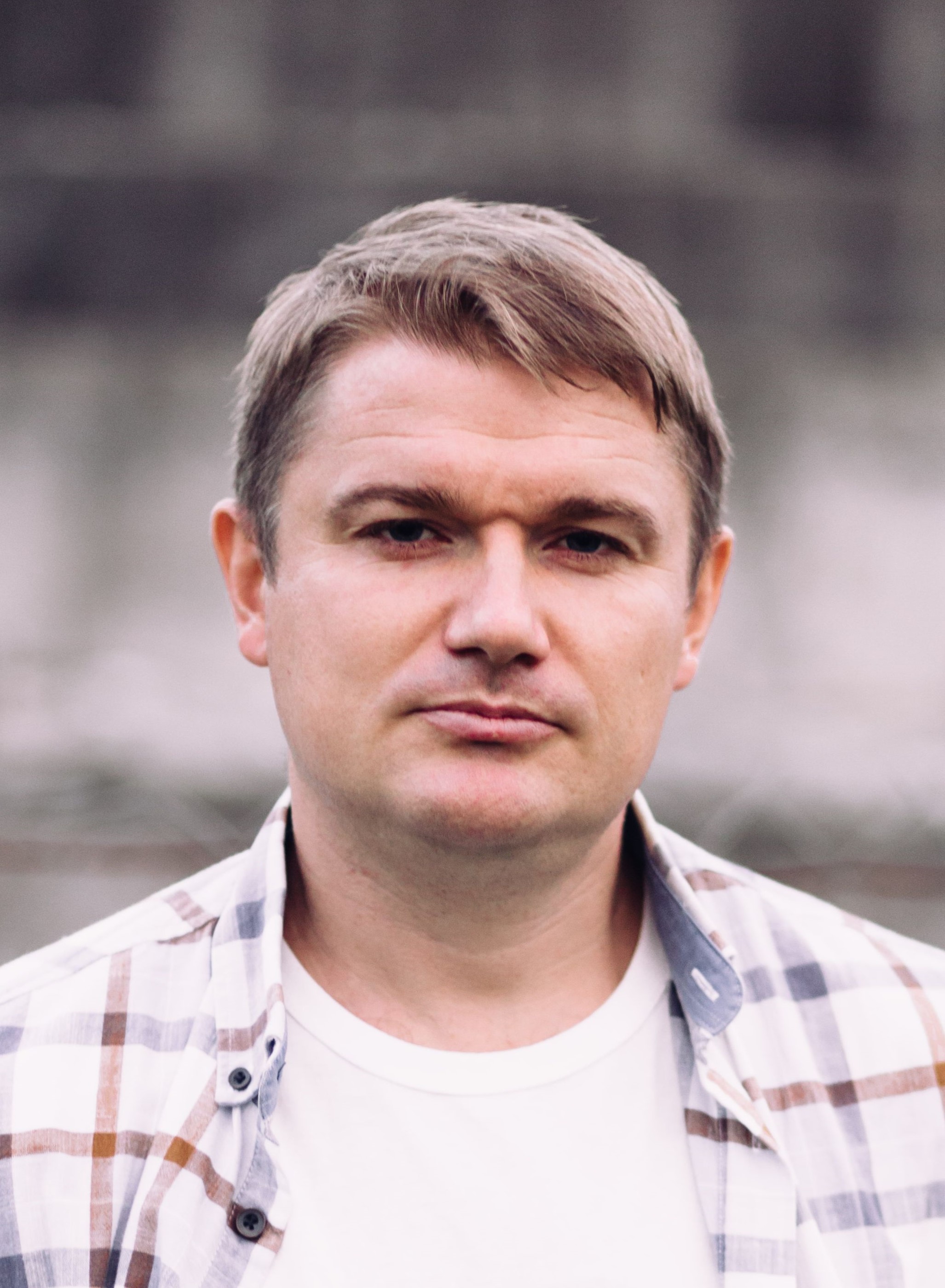 og på engelsk - skriv mann  The image shows a man with short light brown hair, wearing a white t-shirt and an open, light-colored plaid shirt. He is outdoors, and the background is softly blurred and grayish, giving the photo a neutral look. The picture is taken from the chest up.