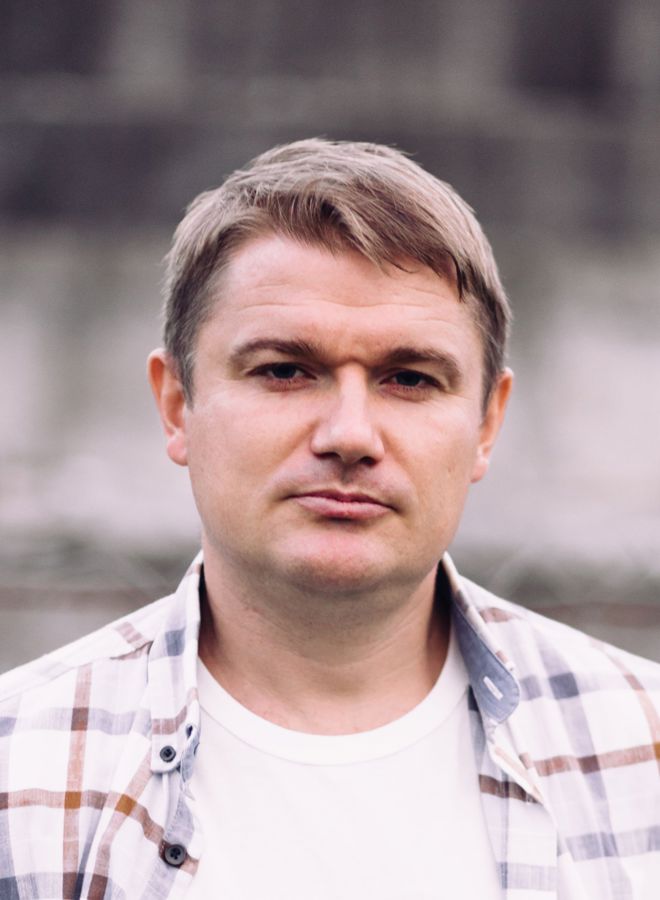 Bildet viser en mann med kort, lysebrunt hår. Vedkommende har på seg en hvit T-skjorte og en rutete skjorte i lyse farger, som er åpen foran.