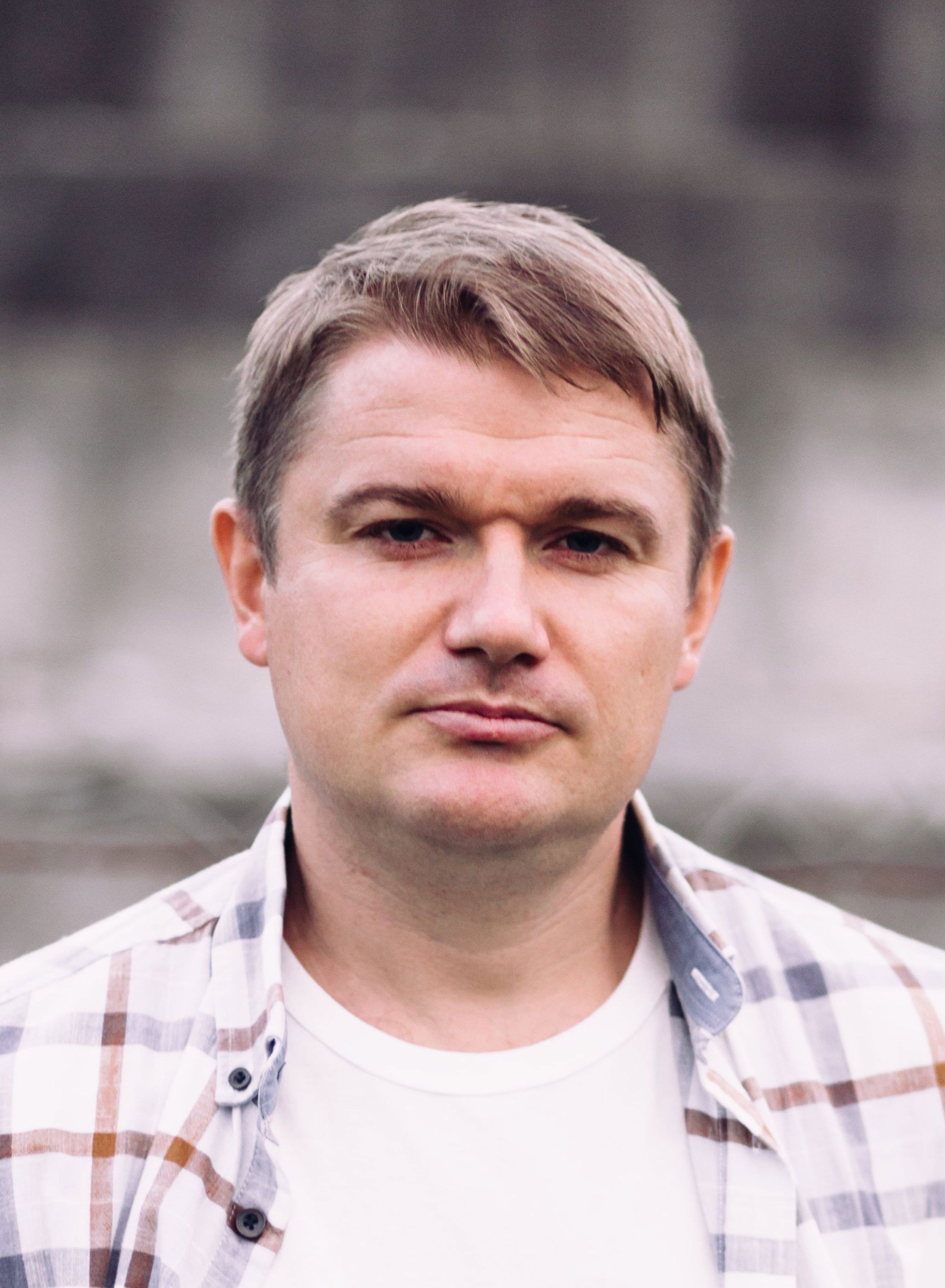 Bildet viser en mann med kort, lysebrunt hår. Vedkommende har på seg en hvit T-skjorte og en rutete skjorte i lyse farger, som er åpen foran.