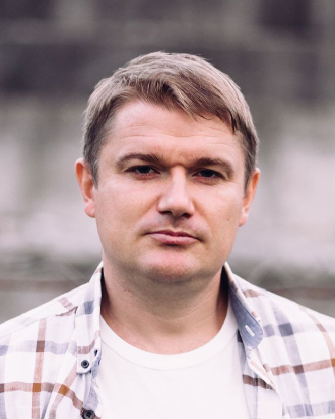og på engelsk - skriv mann The image shows a man with short light brown hair, wearing a white t-shirt and an open, light-colored plaid shirt. He is outdoors, and the background is softly blurred and grayish, giving the photo a neutral look. The picture is taken from the chest up.