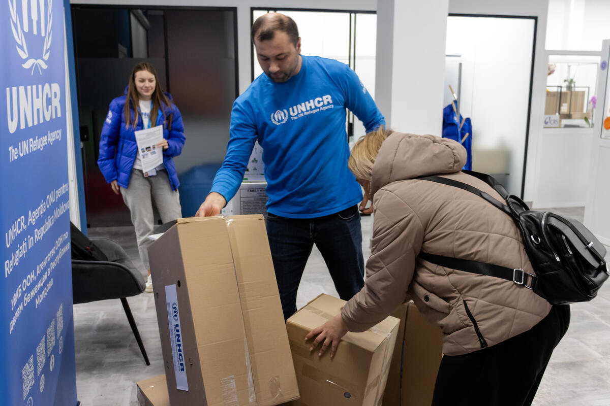 Man wearing a UNHCR sweater helping a woman lift moving boxes.