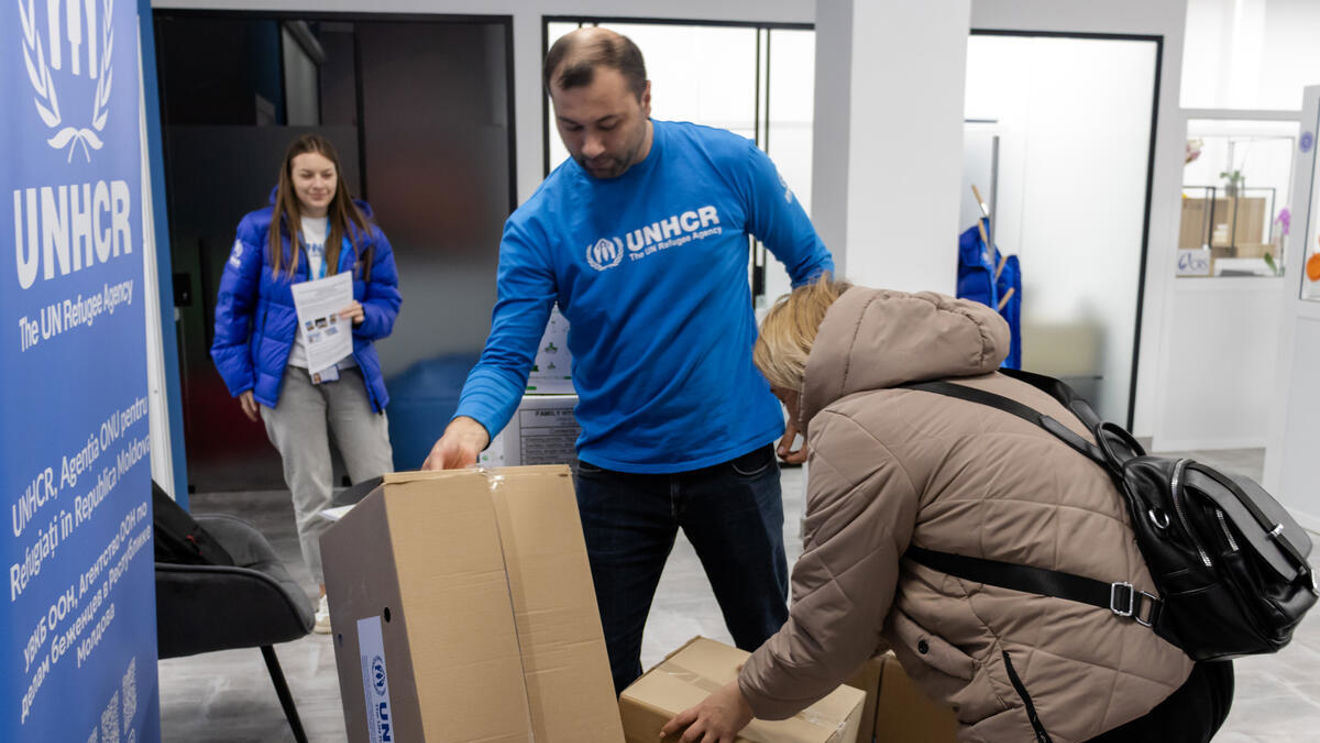 Man wearing a UNHCR sweater helping a woman lift moving boxes.
