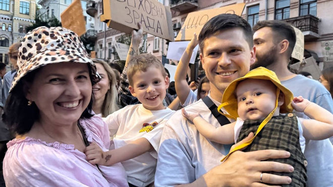 A group of adults and children stand together in a crowd, with some people holding signs in the background.