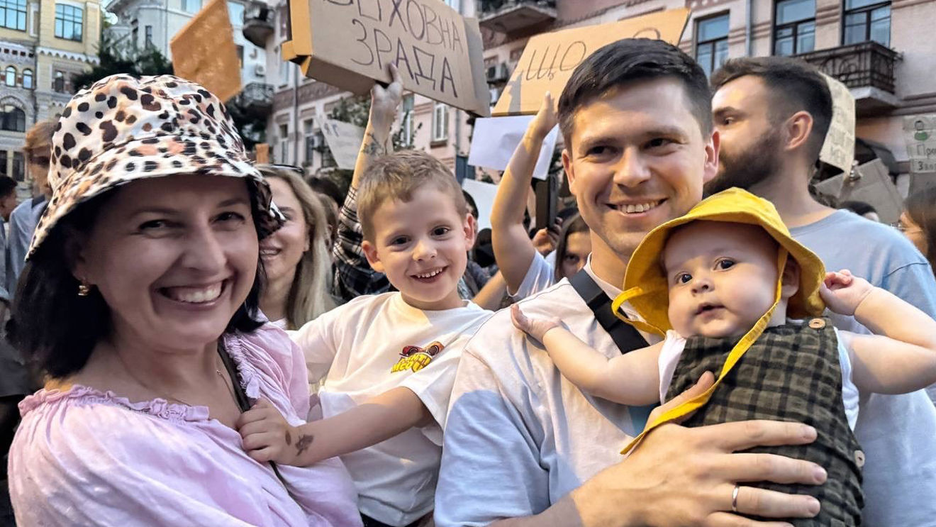 A group of adults and children stand together in a crowd, with some people holding signs in the background.
