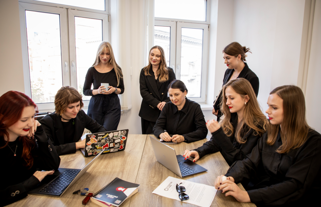 Åtte unge kvinner sitter og står rundt et møtebord med bærbare datamaskiner og papirer, i et lyst rom med store vinduer.