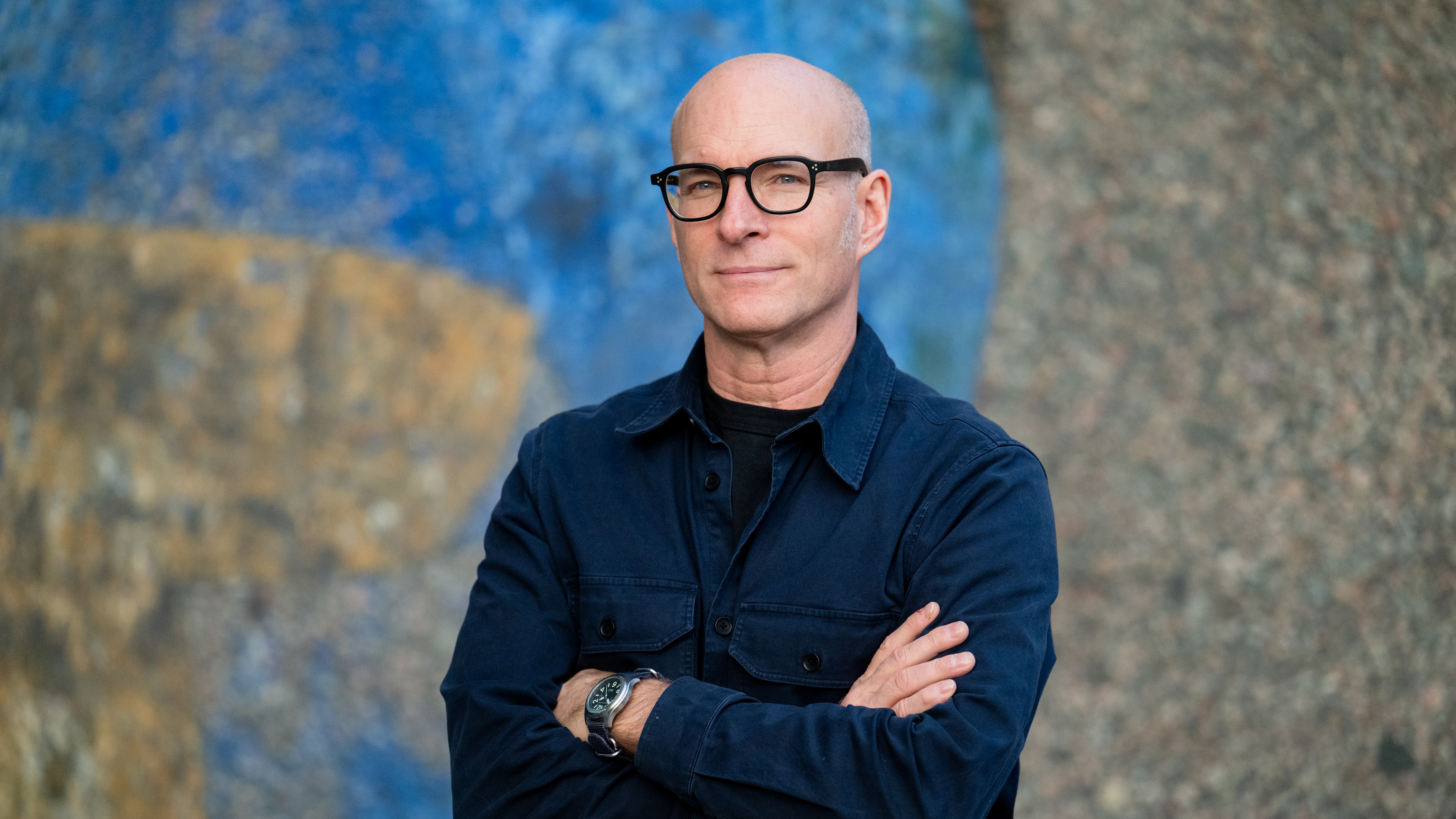 A bald man with glasses stands with arms crossed, wearing a dark jacket in front of a textured, colorful wall.