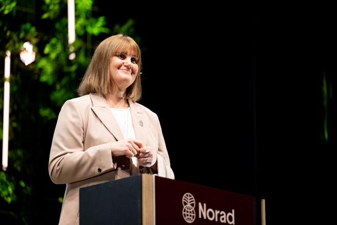 A woman stands at a podium with the Norad logo, wearing a light-colored blazer and speaking at an event.