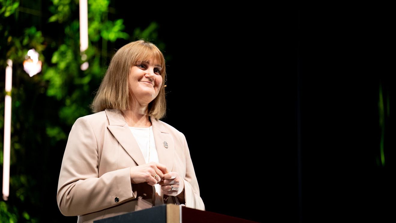 A woman stands at a podium with the Norad logo, wearing a light-colored blazer and speaking at an event.