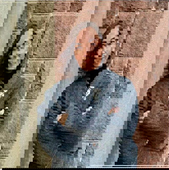 A woman with shoulder-length brown hair stands with arms crossed, wearing a dark coat in front of a brick wall.