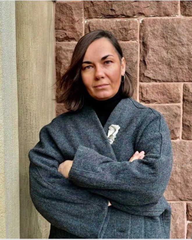 A woman with shoulder-length brown hair stands with arms crossed, wearing a dark coat in front of a brick wall.