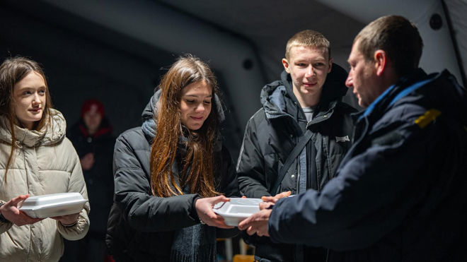 Flere unge personer mottar matbokser fra en voksen i en varm jakke inne i et telt.